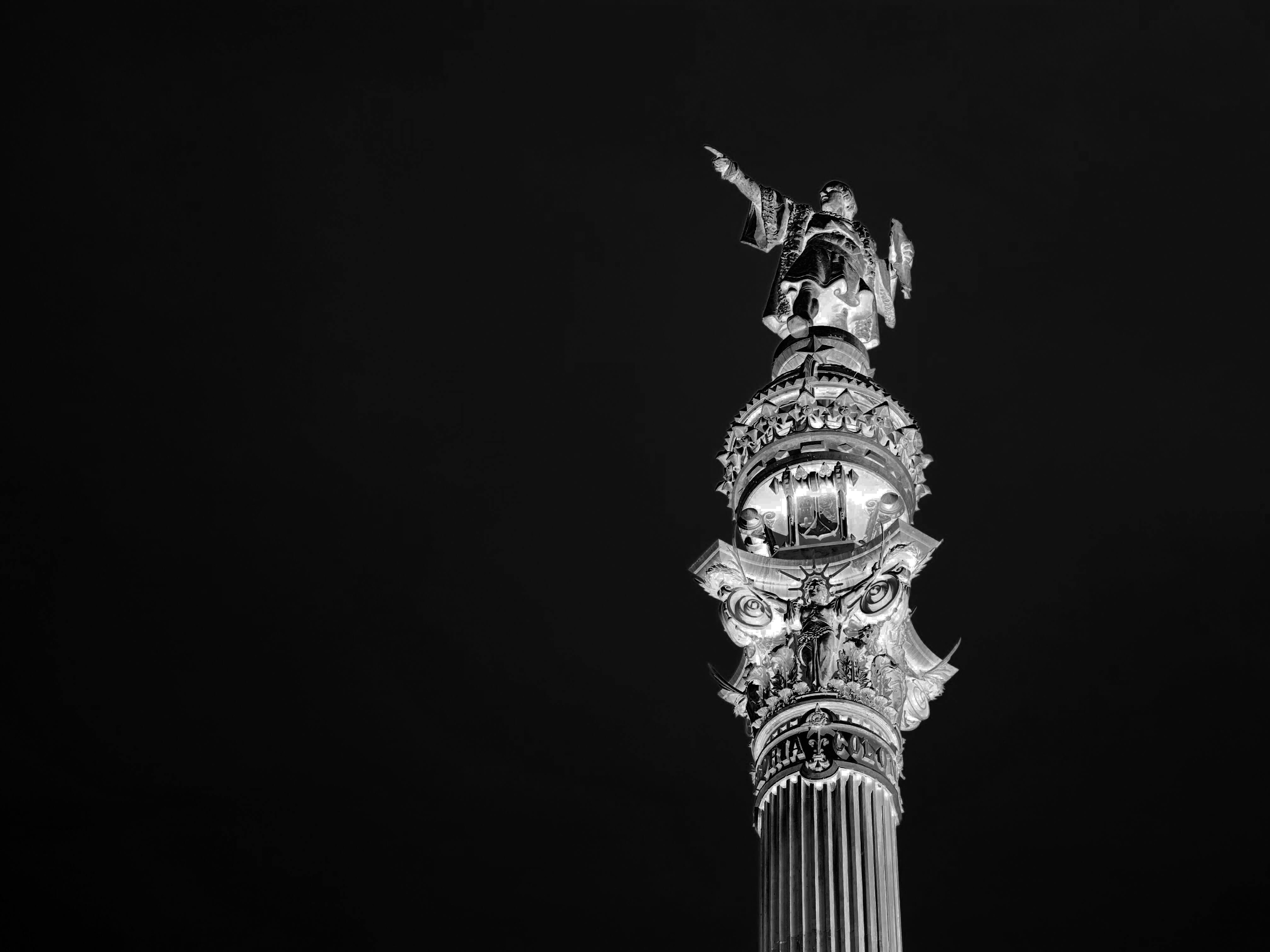 The top of a tall ornate column with a statue pointing outward, lit against a dark sky.