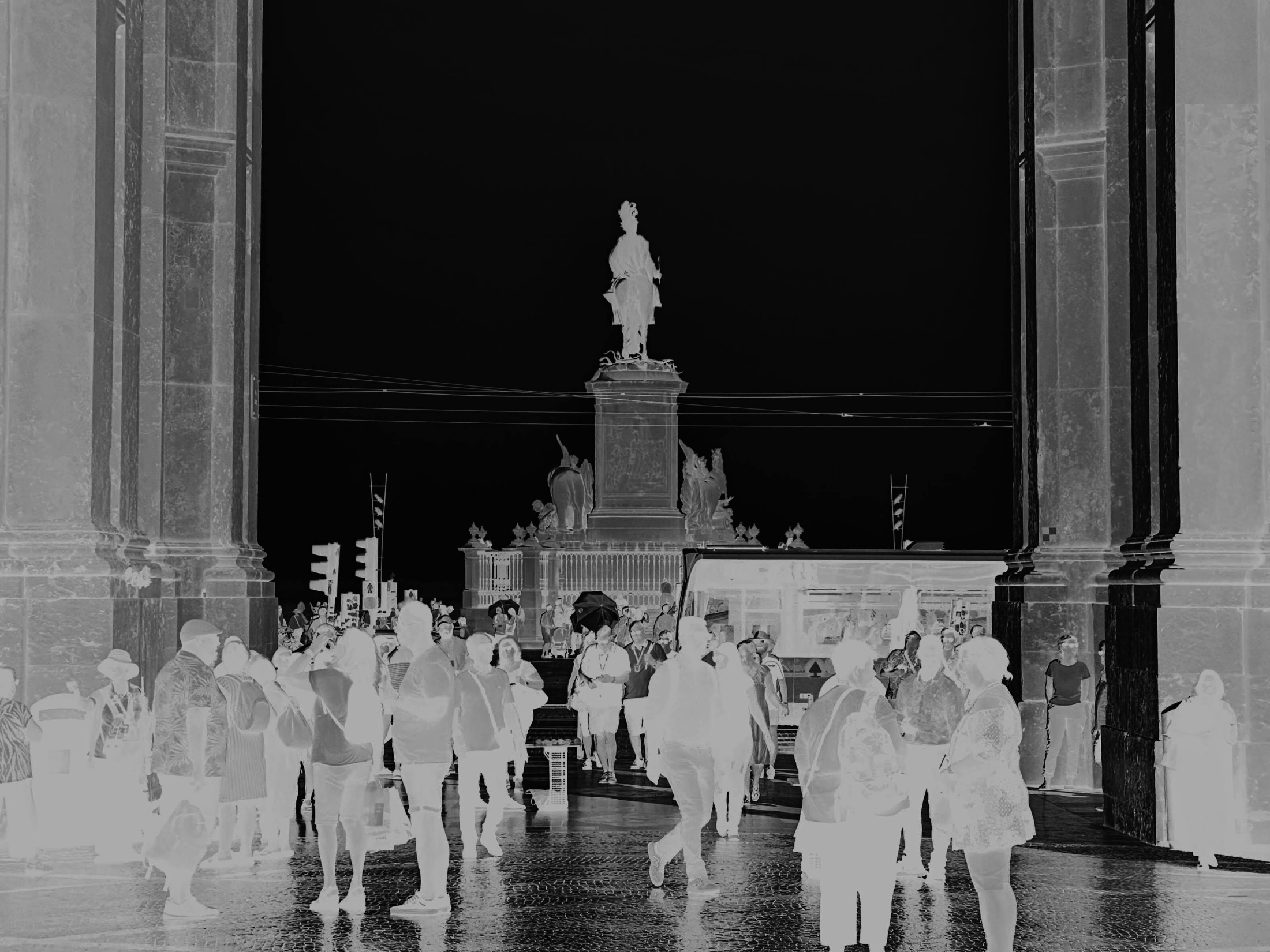 A crowd of people in a plaza framed by large stone columns, with a statue on a pedestal in the background, rendered in negative.